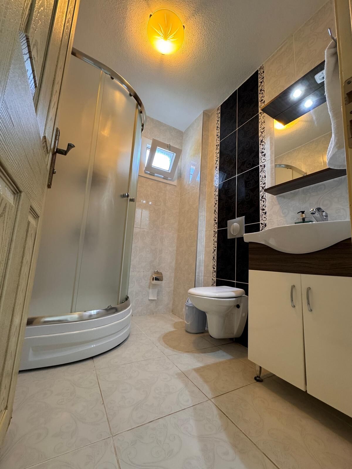 A bathroom with a glass shower enclosure, a toilet, a sink with a mirror, and patterned tiles.