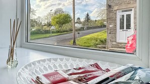 A window with a view of a road and greenery, a glass vase with reeds, and a plate with newspapers. - 2 bedroom accommodation in Stanhope, near Bishop Auckland (Stanhope, near Bishop Auckland)