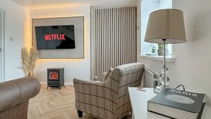 A living room with a flat-screen TV displaying 'NETFLIX', a plaid armchair, a white table with a book titled 'PARIS', and a radiator. - 2 bedroom accommodation in Stanhope, near Bishop Auckland (Stanhope, near Bishop Auckland)