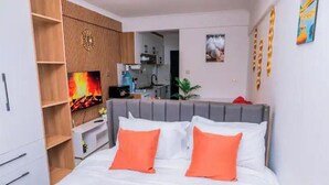 A bedroom with a bed, orange pillows, a wooden wardrobe, a TV, and a kitchen in the background. - Skyview loft retreat Apartment, Smarthome Yaya (Nairobi)