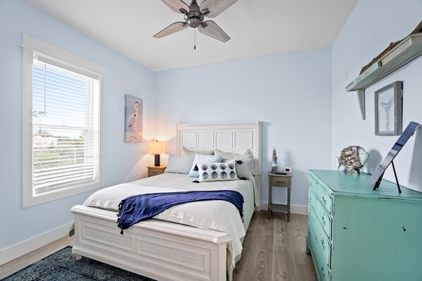Une chambre à coucher avec un lit, un ventilateur de plafond, une commode et une fenêtre avec des stores.
