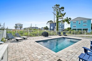 A swimming pool with lounge chairs and a grill, surrounded by a white fence and houses in the background.