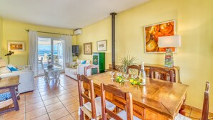 Uma sala de estar iluminada com uma mesa de jantar de madeira, cadeiras e um sofá. Há uma TV na parede e uma janela com cortinas.