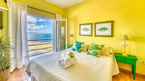 Quarto com cama, mesa de cabeceira verde, luminária, duas fotos emolduradas de peixes e vista para o mar através de uma janela.