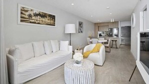 A modern living room with a white sofa, a round coffee table, and a painting of palm trees. - Broadbeach Elegant apartment with views! (Broadbeach)