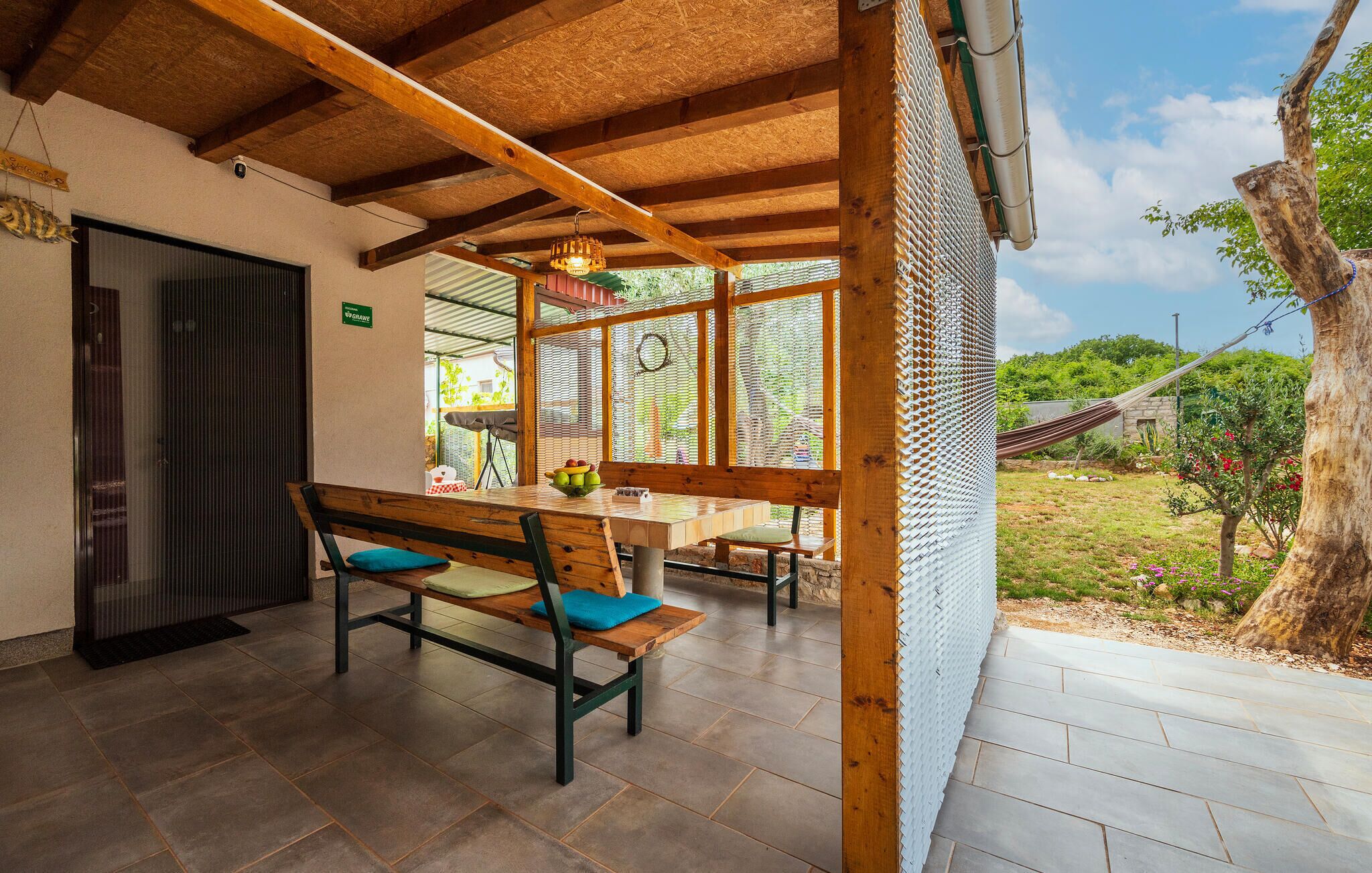 Un espace extérieur abrité, aménagé pour les repas, avec une table et des bancs en bois, une vue sur le jardin et un hamac en arrière-plan.