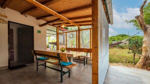 Un espace extérieur abrité, aménagé pour les repas, avec une table et des bancs en bois, une vue sur le jardin et un hamac en arrière-plan.