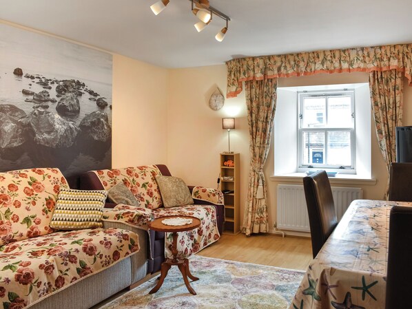 A cozy living room with a floral sofa, a small round table, and a window with curtains. - Cowie Mill Apartment (Stonehaven, near Banchory)