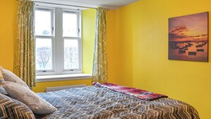 Ein Zimmer mit gelben Wänden, einem Bett mit gemusterten Betttextilien, einem Fenster mit Vorhängen und einem Sonnenuntergangsbild an der Wand.
