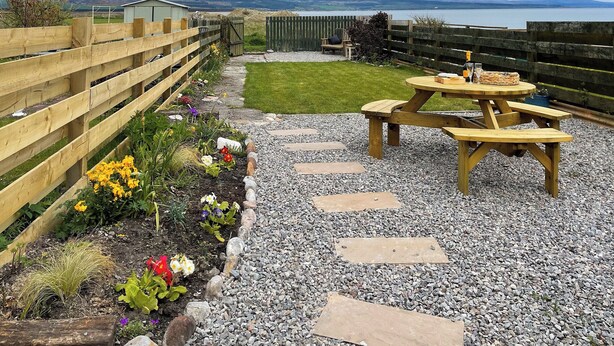 Une terrasse de jardin avec une table et des bancs en bois, entourée d’un chemin de gravier et de parterres de fleurs.