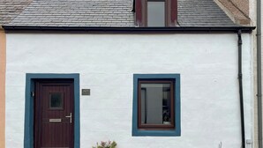 Un bâtiment blanc avec une porte et une fenêtre de couleur brun foncé, une petite fenêtre de toit et un pot de fleurs à sa base.