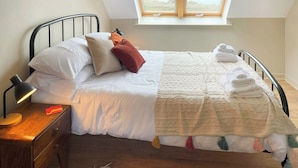 Un lit bien fait, avec du linge de lit blanc et une couverture beige, une lampe de chevet et une table de chevet en bois dotée d’un tiroir.