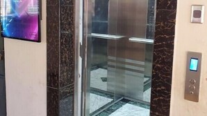 Un ascensor con pantalla digital y puerta de vidrio, rodeado de paredes de mármol y suelo de baldosas.