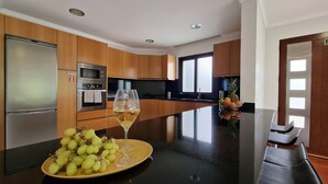 A modern kitchen with stainless steel appliances, wooden cabinets, and a central island with a glass of white wine and a bowl of green grapes. - Capelas House by LovelyStay (Campanario)