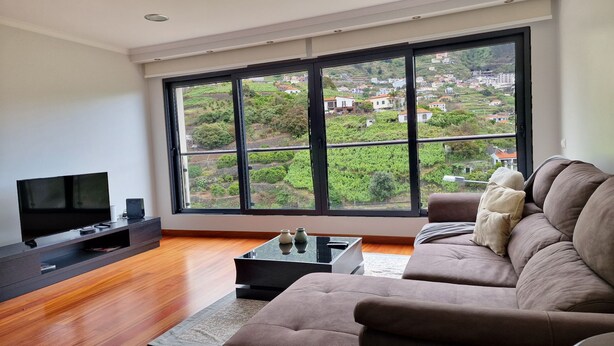 A modern living room with a large window, a flat-screen TV, a sofa, and a coffee table. - Capelas House by LovelyStay (Campanario)