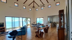A spacious living area with a large window overlooking the ocean, a dining area with a wooden table, and a kitchen in the background. - Casa Celajes (Chomes)