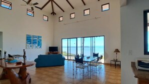 A spacious living room with a large window overlooking the sea, a blue sofa, a dining table, and a TV. - Casa Celajes (Chomes)