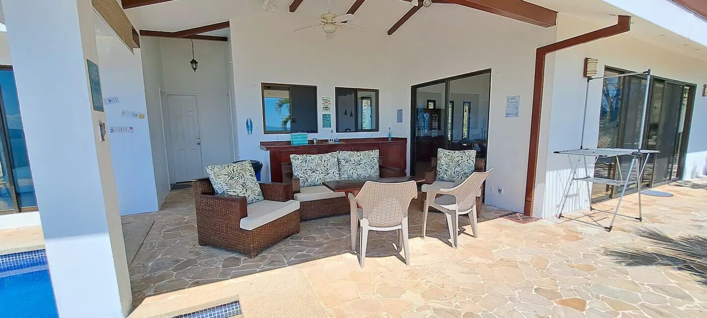 A covered outdoor seating area with wicker furniture and a stone floor, leading to a pool.