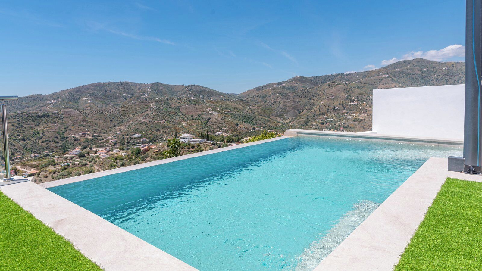 Une piscine rectangulaire à l’eau d’un bleu limpide, entourée d’une terrasse en béton et d’une pelouse. Au loin, on aperçoit des collines parsemées de bâtiments.