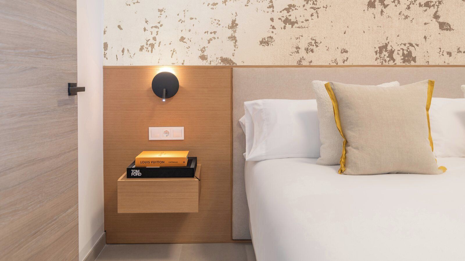 A hotel room with a wooden headboard, a bedside table with a Louis Vuitton and Tom Ford box, and a wall-mounted light.