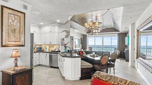 A spacious kitchen with white cabinets, a central island, and a dining area with a view of the ocean. - Wave Whisperer: Family Beachfront Hideaway (Destin)