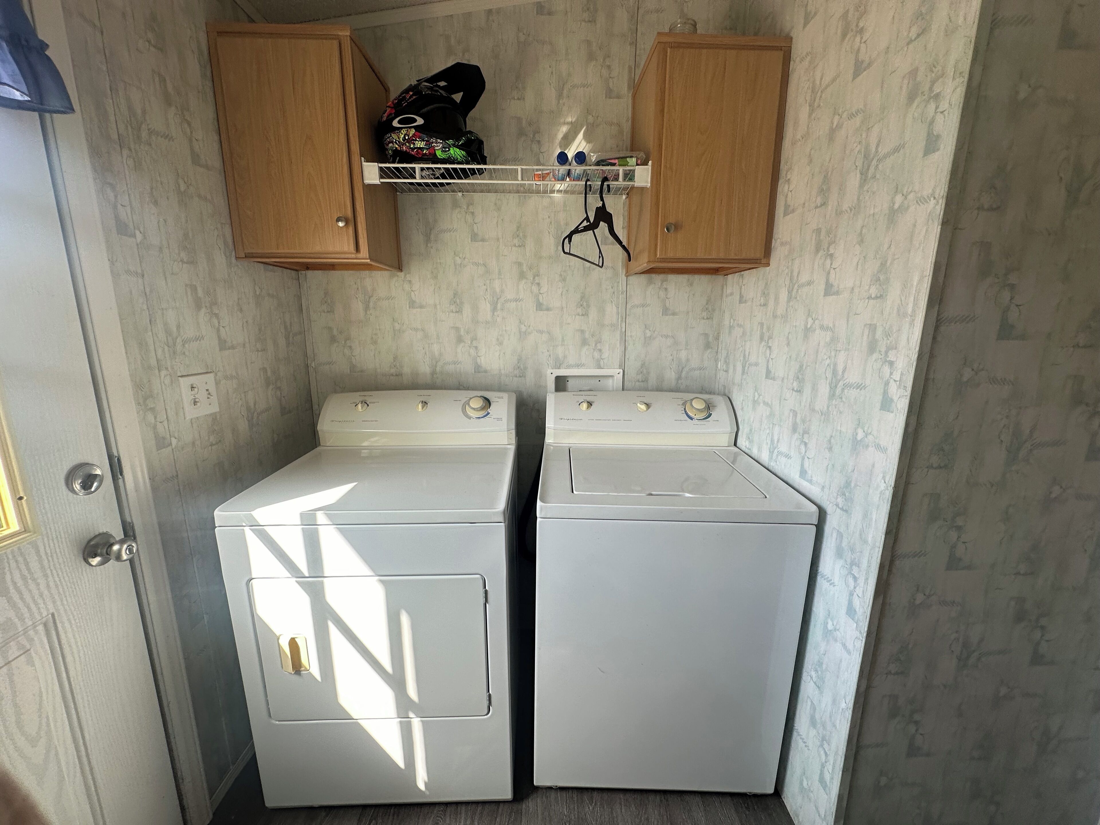 A compact laundry room with two white front-loading washers and dryers, wooden cabinets above, and a small shelf with personal items.