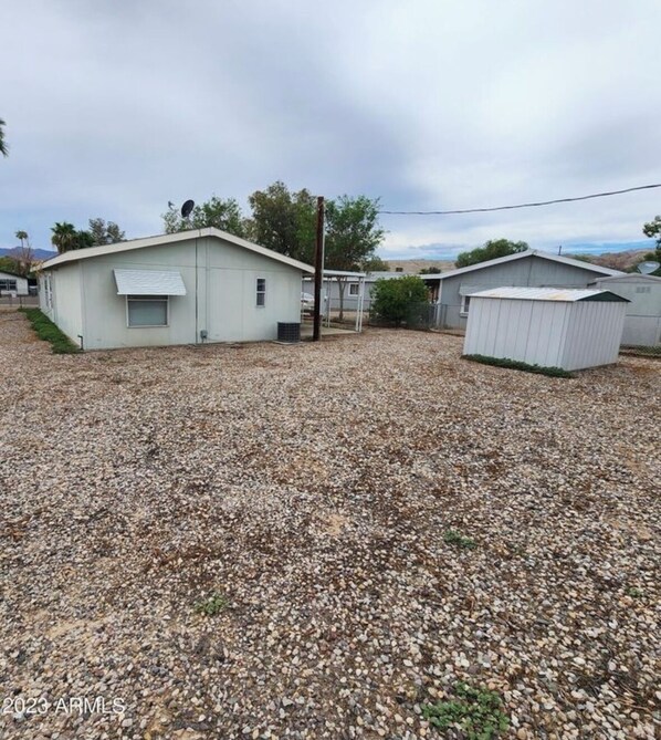 A gravel-covered yard with two small, single-story buildings, one with a slanted roof and a small window, and the other with a flat roof and a door.