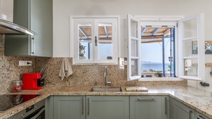 A kitchen with granite countertops, a red coffee maker, a built-in oven, and a view of the sea through a window. - Holiday Home 'Waterfront Masterpiece' with Sea View, Wi-Fi and Air Conditioning (Tinos)