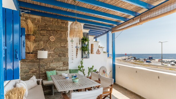 A covered outdoor seating area with a stone wall, a table, chairs, and a view of the sea. - Holiday Home 'Waterfront Masterpiece' with Sea View, Wi-Fi and Air Conditioning (Tinos)
