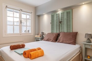 A neatly made bed with rolled towels, a bedside table with a lamp, and a window with shutters.