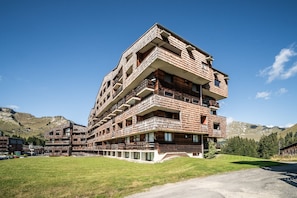 Un hôtel à plusieurs étages doté de balcons, d’une allée gravillonnée et surplombant un paysage montagneux.