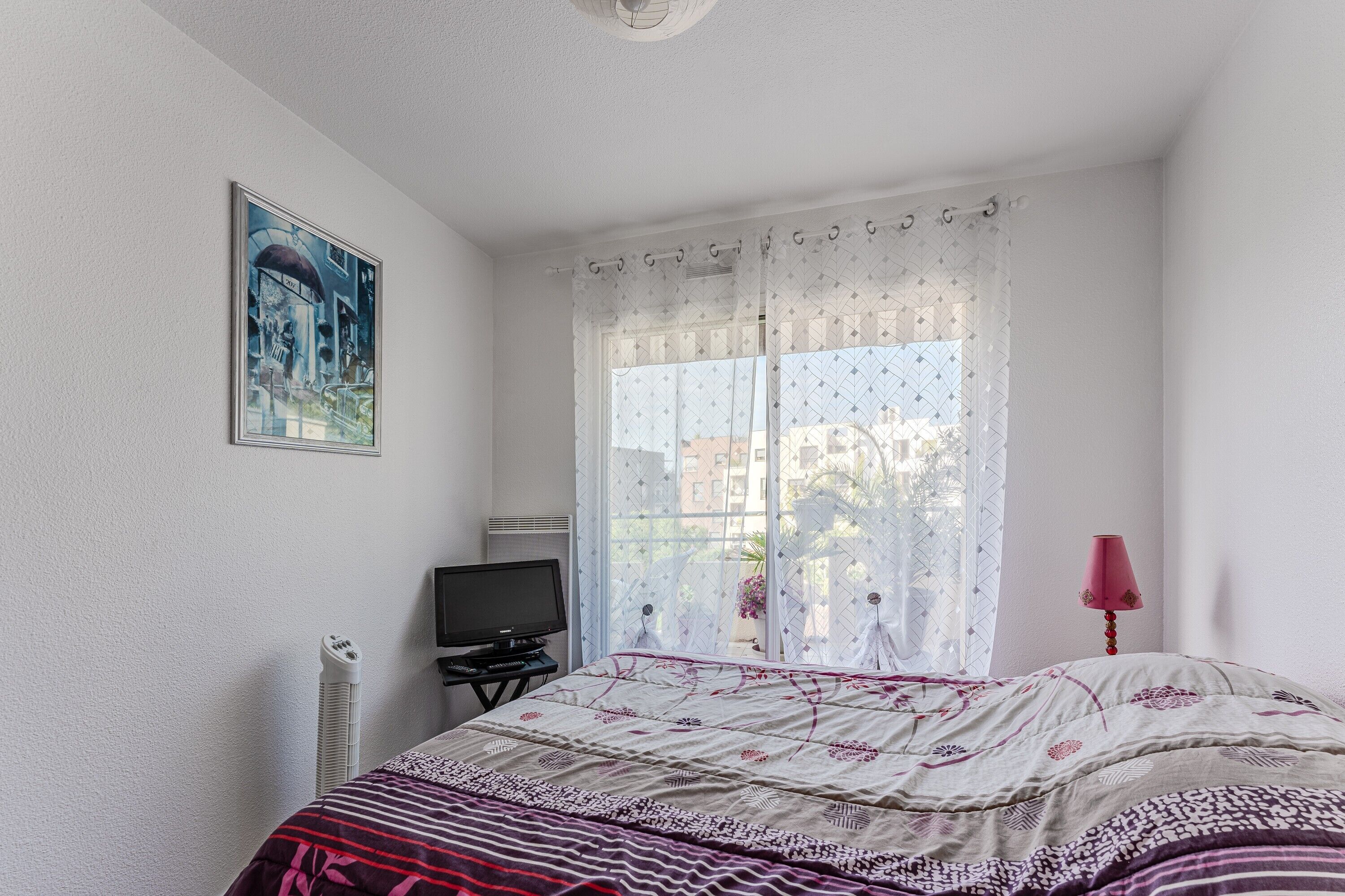 Une chambre à coucher avec un lit, une télévision, un radiateur et une fenêtre avec un rideau.