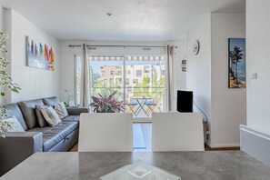 A modern living room with a grey sofa, a glass coffee table, a balcony with a view of buildings and trees, and a painting on the wall.