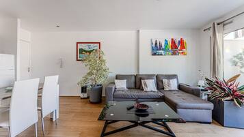 Een moderne woonkamer met een bank, een eettafel met stoelen, een salontafel, een potplant en een kleurrijk schilderij aan de muur.