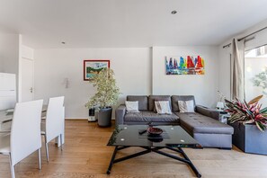 A modern living room with a sofa, a dining table with chairs, a coffee table, a potted plant, and a colorful painting on the wall.