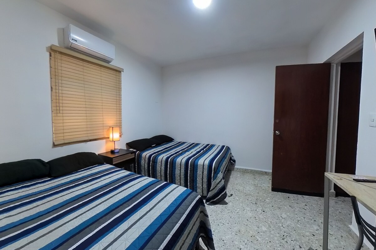 A room with two beds, a desk, a chair, a lamp, a window with blinds, an air conditioner, and a door.
