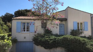 A two-story building with a tiled roof, a garage door, and a tree with pink flowers. - Holiday Home 'Jardin Arboré, Belle Maison Piscine' with Private Pool, Terrace and Wi-Fi (Saint-Trojan-les-Bains)