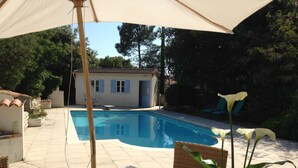 A swimming pool with a tiled deck, a covered patio area, and a building with blue shutters in the background. - Holiday Home 'Jardin Arboré, Belle Maison Piscine' with Private Pool, Terrace and Wi-Fi (Saint-Trojan-les-Bains)