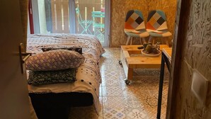 A bedroom with a bed, a small table, two chairs, a sliding glass door to a balcony, and a wall with potted plants.