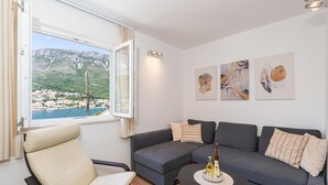 Un salon moderne avec un canapé d’angle gris, un fauteuil beige et une table basse ronde sur laquelle se trouvent une bouteille et des verres. Une fenêtre offre une vue sur une ville côtière et des montagnes.