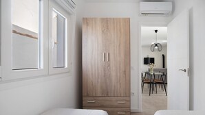 A bedroom with two beds, a wardrobe, a window, and a dining area in the background.