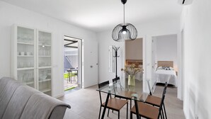 A modern living room with a glass dining table, a sofa, and a kitchen area with cabinets and a hanging light fixture.