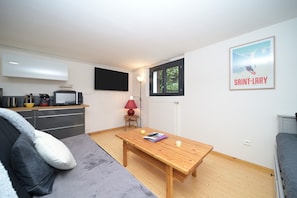 A small, modern room with a wooden table, a grey sofa, a TV mounted on the wall, and a poster on the wall.