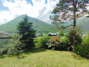 A grassy area with a mountain in the background, a few trees, and a building with a sloped roof.