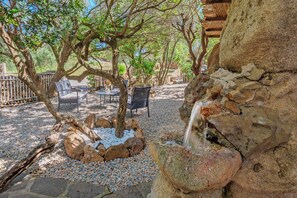 Un área de estar al aire libre tranquila, con mesas y sillas, una pequeña fuente de agua y una rústica pared de piedra.