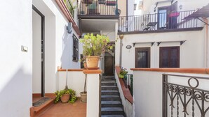 A narrow alleyway with a staircase leading to a second-floor balcony, potted plants, and a metal railing.