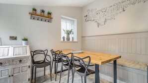 Ein Essbereich mit einem Holztisch, vier Stühlen, einer Zierkommode, einem an der Wand befestigten Regal mit Pflanzen, einem Fenster mit Jalousien und einem Wandgemälde.