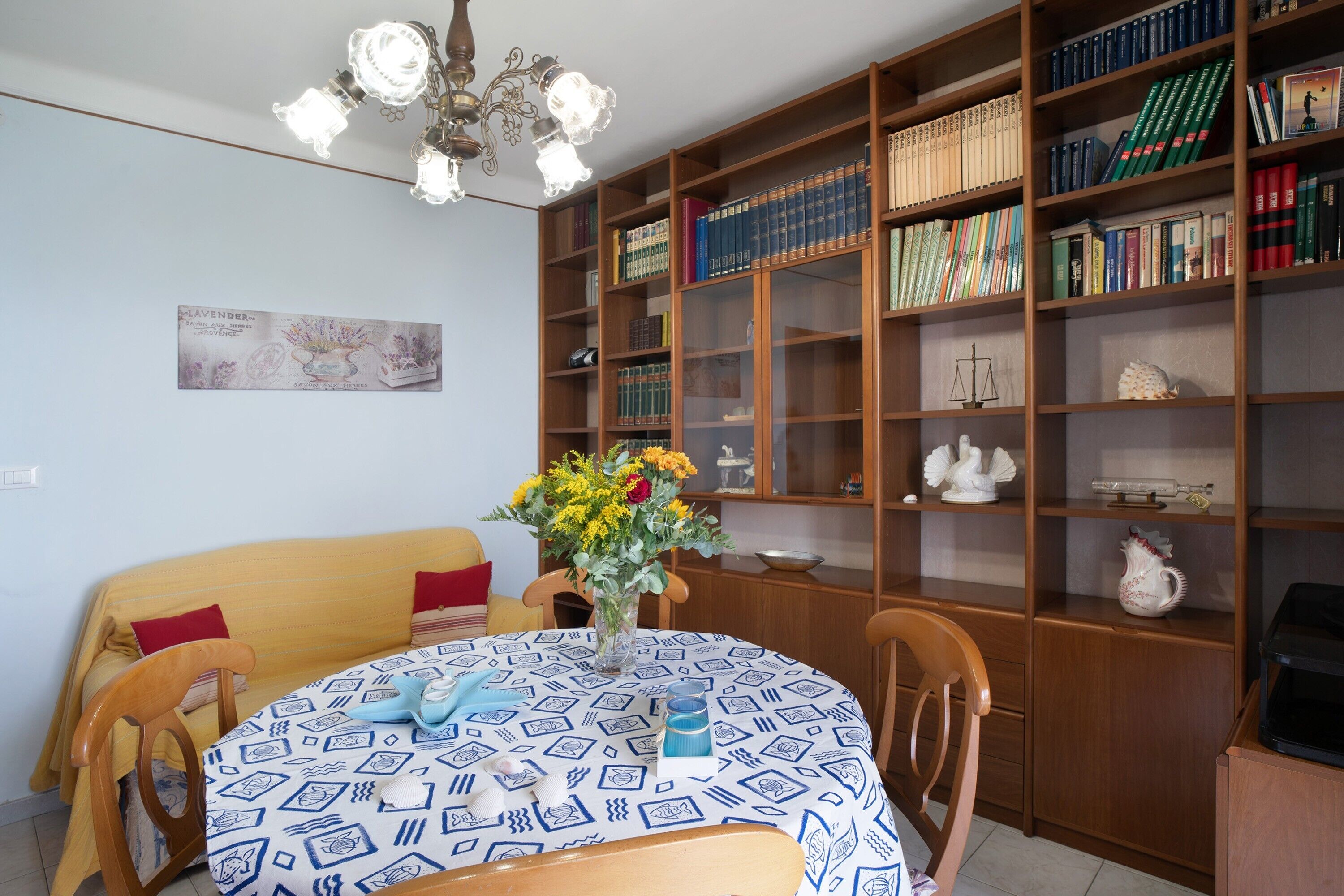 A room with a round table, chairs, a bookshelf, and a chandelier.