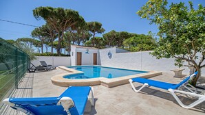 Una piscina con sillas reclinables azules, un edificio pequeño con una puerta y árboles al fondo.