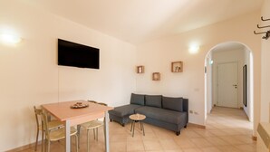 Una sala de estar con sofá, una mesa de comedor con sillas, un televisor de pantalla plana montado en la pared y una mesita auxiliar.